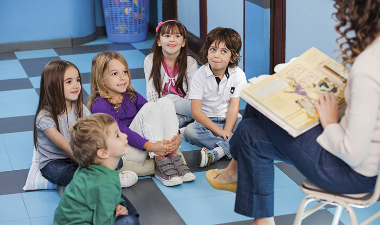 Professora sentada em uma cadeira branca, com um livro colorido e ilustrado aberto em seu colo. A sua frente estão cinco crianças sorridentes, três meninas e dois meninos, de aproximadamente 7 anos, sentadas no chão quadriculado cinza e azul da sala de aula.